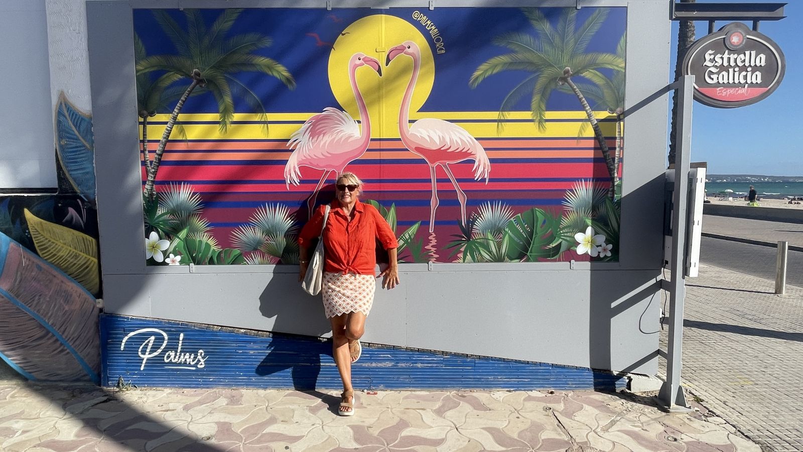 „Vor einem farbenfrohen Plakat auf Mallorca steht Claudia und lächelt in die Kamera.“