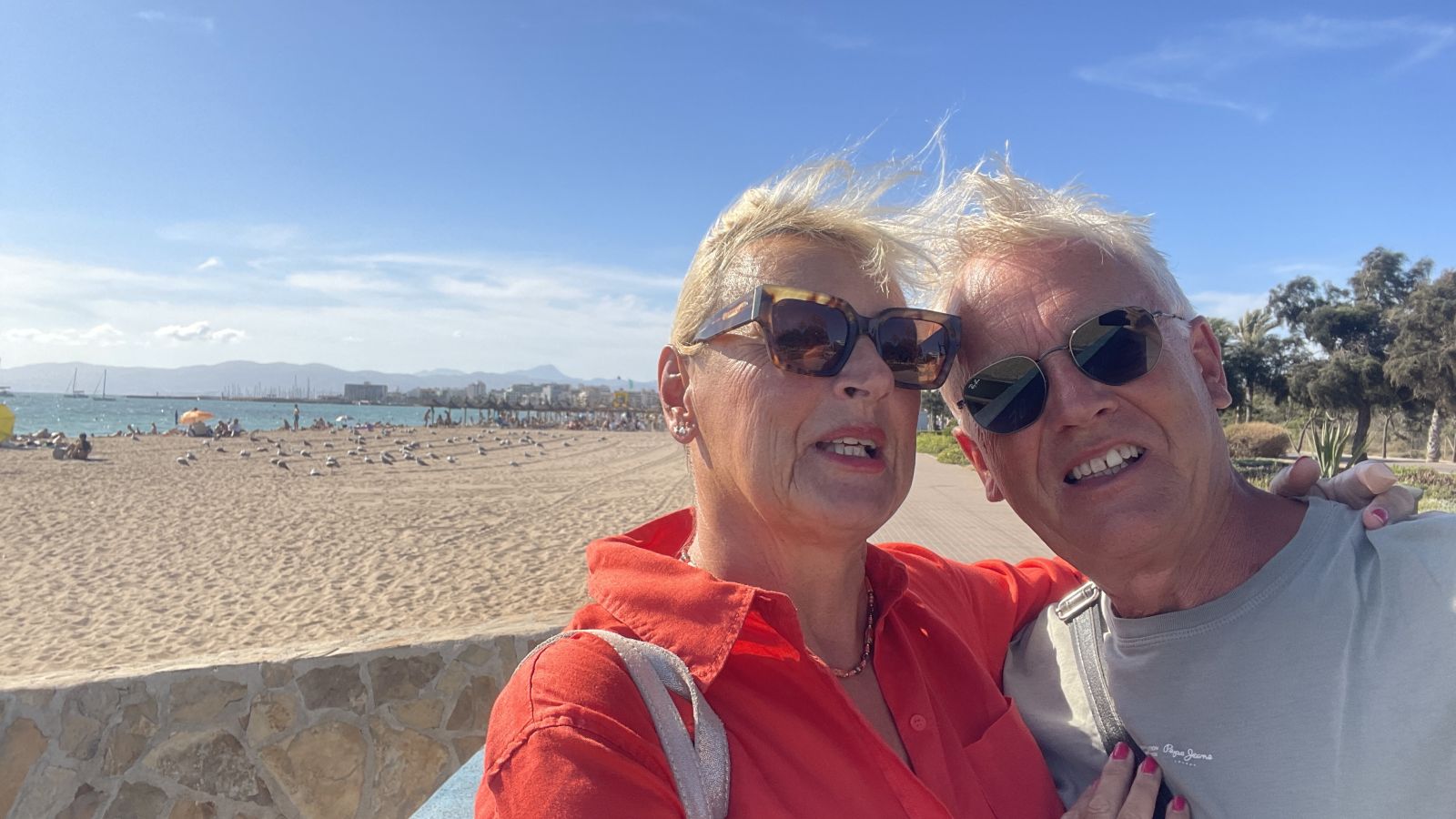 Axel und Claudia spazieren gemeinsam am Strand von Mallorca, genießen die Meeresbrise und den Sonnenuntergang.